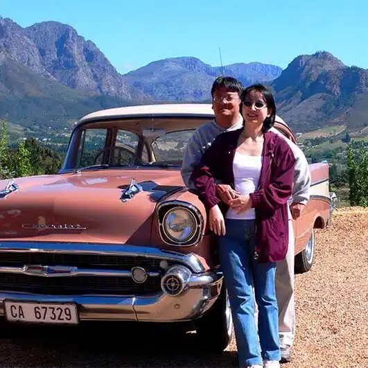 Couple standing in front of a classic pink car with mountains behind, ready for a vintage car transfer Couple standing in front of a classic pink car with mountains behind, ready for a vintage car transfer